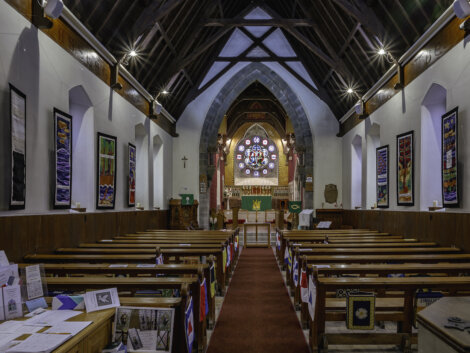 Interior of a small church with wooden pews, colorful banners hanging on the walls, an arched ceiling, and a stained glass window near the altar.