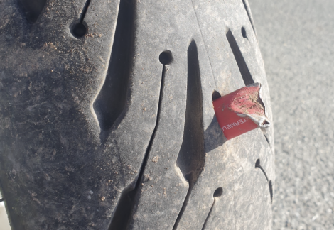 A close-up image of a tire with a sharp metal piece embedded in the tread, causing visible damage. The tire has a textured surface, and the background shows a paved road.