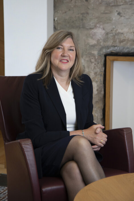 A seated woman wearing a dark blazer and white blouse, with hands resting on her lap, in an office setting with a textured wall background.