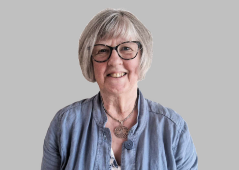 A woman with short gray hair and glasses is smiling. She is wearing a blue top and a necklace with a circular pendant. The background is gray.