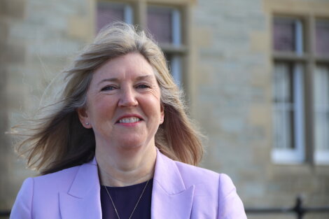 A woman with long blond hair smiles, wearing a light purple blazer, standing in front of a building with stone walls and windows.