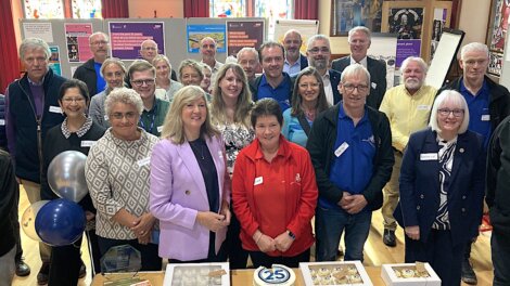 A group of people posing for a photo at an indoor event. Tables with a 25th anniversary cake, cupcakes, and balloons are in the foreground. Poster boards with information are visible in the background.