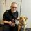 A man in uniform holds a medal while kneeling beside a Labrador retriever wearing a harness. They both face the camera inside an indoor setting.