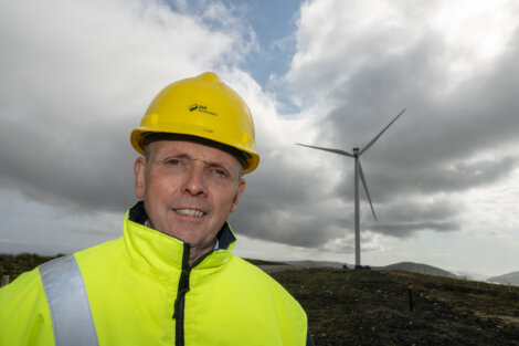 A man in a yellow hard hat and high-visibility jacket stands in a hilly area with a wind turbine in the background under a cloudy sky.