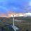 A wind farm with multiple wind turbines stands on a vast, hilly landscape under a partly cloudy sky at sunset.
