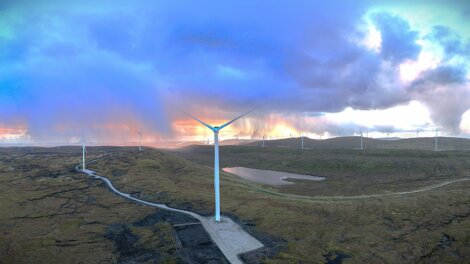 A wind farm with multiple wind turbines stands on a vast, hilly landscape under a partly cloudy sky at sunset.