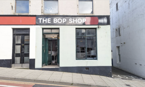 Street view of "The Bop Shop," a storefront with large windows and a black sign with white text above the entrance door. The surrounding buildings are white and grey.