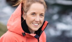 A woman with light brown hair tied up, wearing an orange hooded jacket, smiles at the camera with a blurred outdoor background.