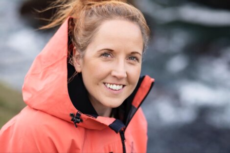 A woman with light brown hair tied up, wearing an orange hooded jacket, smiles at the camera with a blurred outdoor background.