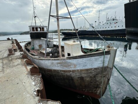 A weathered, wooden fishing boat tied to a concrete pier with a larger vessel in the background on a cloudy day.