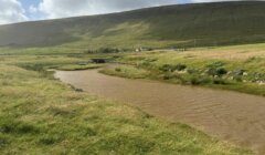 A muddy river flows through a grassy valley with a green hill in the background under a cloudy sky. A small bridge and scattered buildings are visible in the distance.