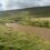 A muddy river flows through a grassy valley with a green hill in the background under a cloudy sky. A small bridge and scattered buildings are visible in the distance.
