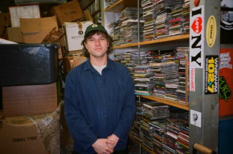 Person standing in a cluttered room, surrounded by stacks of CDs and boxes. The individual is wearing a blue jacket. Shelving units are seen in the background with more CDs and miscellaneous items.