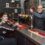 Two people are in a bar. The woman is seated at the bar counter, and the man is standing behind it, with beer taps visible in front of them.