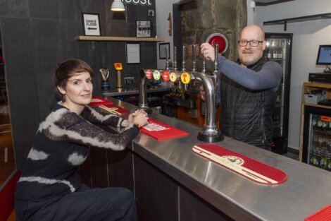 Two people are in a bar. The woman is seated at the bar counter, and the man is standing behind it, with beer taps visible in front of them.