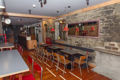 A restaurant interior with wooden flooring, brick walls, and a mix of black and wooden chairs and tables. Red pendant lights hang from the ceiling. The wall features a large mirror and no patrons are present.