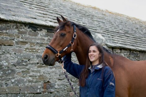A person standing beside a brown horse, holding its reins, with a stone building in the background.