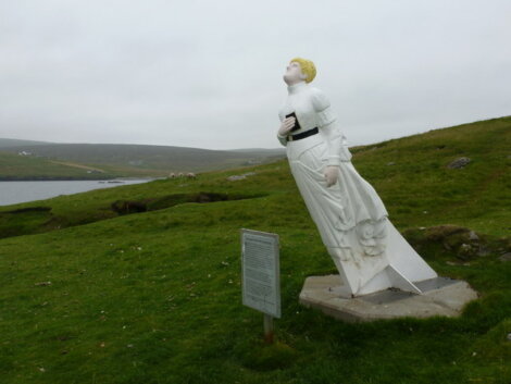 A statue of a woman in a white dress, leaning into the wind, stands on a grassy hill with a sign nearby, overlooking water and distant hills.