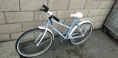 A white and blue bicycle is parked next to a brick wall on a paved surface.