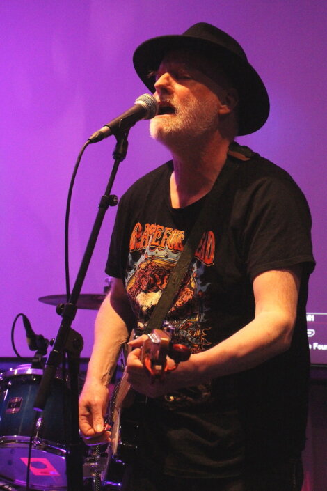 A bearded man in a black hat and graphic T-shirt plays an electric guitar and sings into a microphone on stage. A drum set is visible in the background under purple lighting.