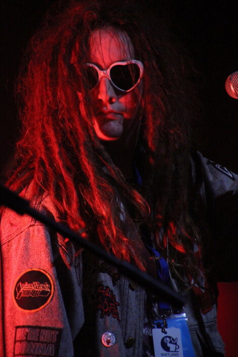 A person with long, dreadlocked hair wears white sunglasses and a jacket with patches, standing in front of a microphone. Red lighting gives the scene a dramatic effect.