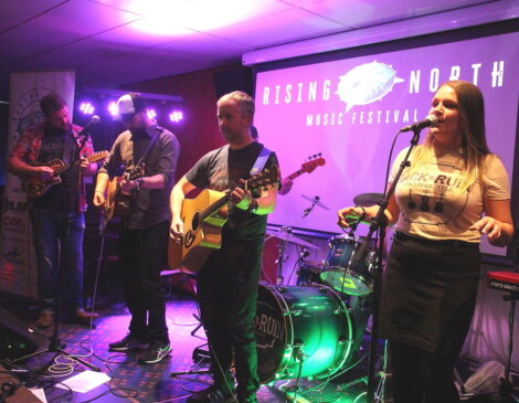A band with three guitarists and a drummer performs on stage at the Rising North Music Festival. One of the guitarists and a singer/bassist are in the foreground.