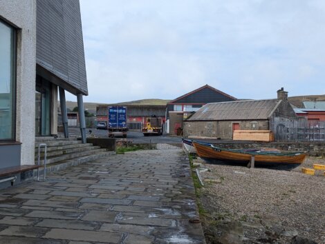 Waterside scene depicting a stone walkway, a docked wooden boat, and an industrial area with blue shipping containers and a forklift in the background.