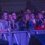 A group of people stand at a barricade, watching an event. Some are holding drinks, and others are taking photos. The area is dimly lit with purple lighting.