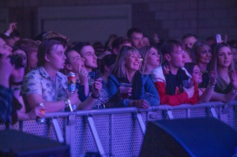 A group of people stand at a barricade, watching an event. Some are holding drinks, and others are taking photos. The area is dimly lit with purple lighting.