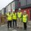 Four people wearing yellow safety vests and white helmets stand in front of newly constructed homes.