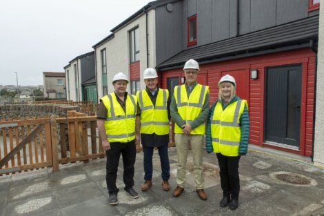 Four people wearing yellow safety vests and white helmets stand in front of newly constructed homes.