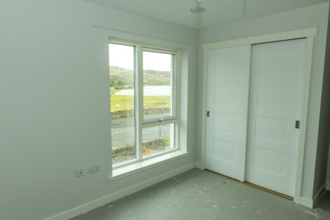 A small, empty room with a window showing an outdoor landscape, and white sliding closet doors. Walls and ceiling are white, flooring is unfinished.