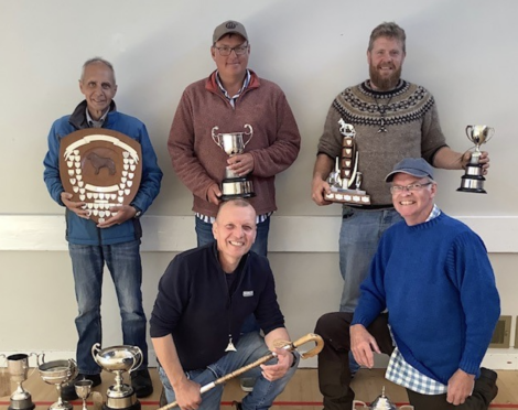 Five smiling men pose with various trophies and awards, standing against a plain background. The group is proudly displaying their achievements, including shields and cups.
