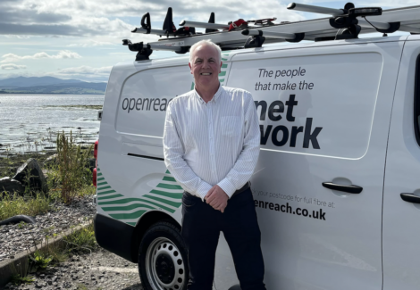 A man stands in front of a white van with "Openreach" branding. The van is parked on a gravel area near a large body of water.