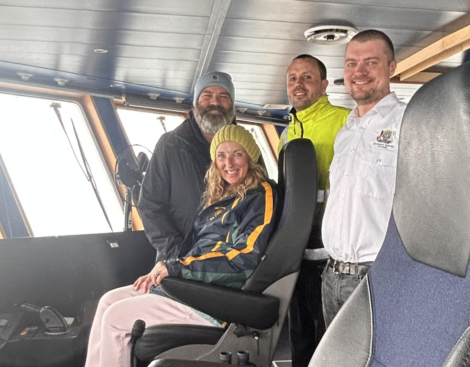 Four people onboard a boat's bridge, smiling at the camera. One person is seated while the others stand behind. The background shows the interior of the bridge with large windows.