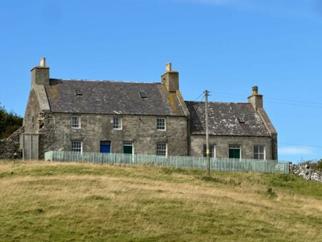 A stone house with two chimneys and a slate roof stands on a grassy hill, surrounded by a light blue picket fence under a clear blue sky.