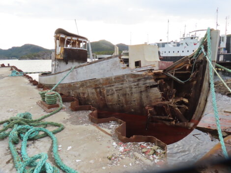 An old, damaged wooden boat is docked at a pier. The hull appears heavily deteriorated, with visible rot and missing planks. Green ropes are strewn around the dock.
