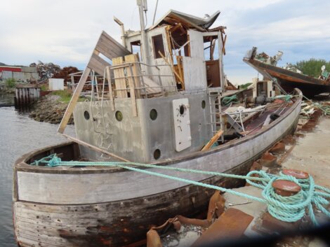 An old, dilapidated boat sits partially submerged and damaged at a dock, with debris and another damaged vessel nearby.