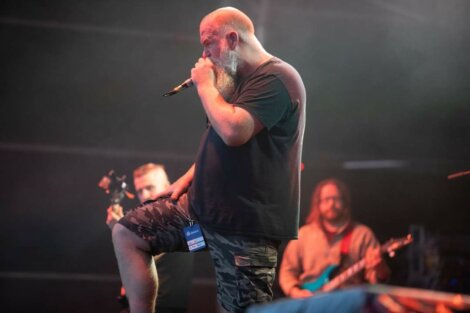A man with a beard and microphone sings on stage, wearing a black shirt and camouflage shorts. Two other musicians are visible in the background performing.