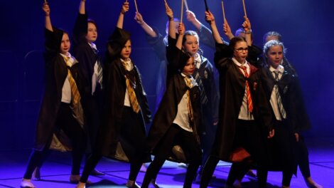 Group of children in wizard costumes holding wands on stage, performing in front of a dark, smoky background.