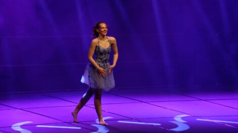 A dancer in a purple and blue costume stands center stage under purple lighting, smiling and appearing to finish a performance.