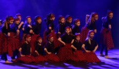 A group of children in matching black shirts and red-striped skirts perform a choreographed dance on stage under purple lighting.