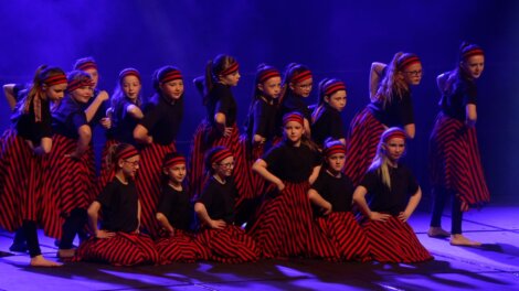 A group of children in matching black shirts and red-striped skirts perform a choreographed dance on stage under purple lighting.