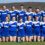 A sports team in blue and white uniforms poses on a field with a trophy, standing in two rows with a coach on the left and another individual on the right.