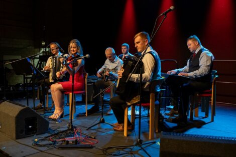 A group of six musicians perform on stage with instruments including accordions, a violin, a keyboard, and a guitar. They are seated under red stage lighting with microphones in front.