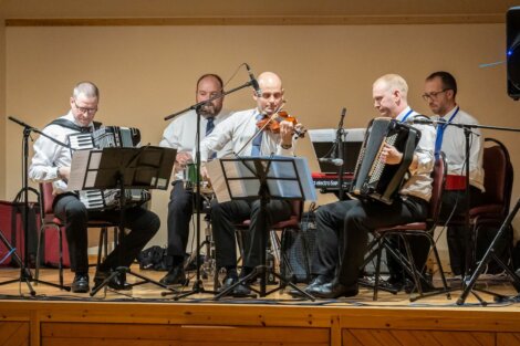 A quintet of musicians dressed in formal attire perform on stage with accordions, a violin, and percussion instruments.