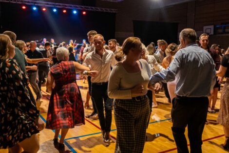 A group of people are dancing on a gymnasium floor under stage lights, engaging in what appears to be a social gathering or dance event.