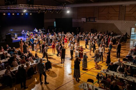 A crowd of people, some seated at tables and others dancing, gather in a gymnasium with a stage setup for a live performance in the background.