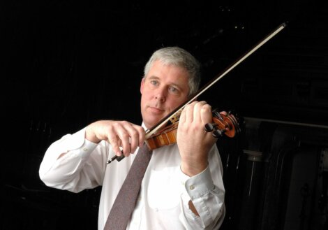 A man in a white shirt and tie plays a violin in a dark setting.