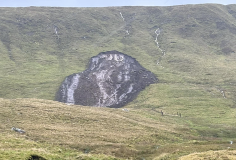 A large exposed section of soil or rock on a grassy hillside, likely caused by erosion or a landslide, with several smaller erosion marks visible on the surrounding slope.
